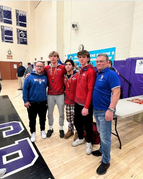 Three high school wrestlers stand with their coaches in a gym.