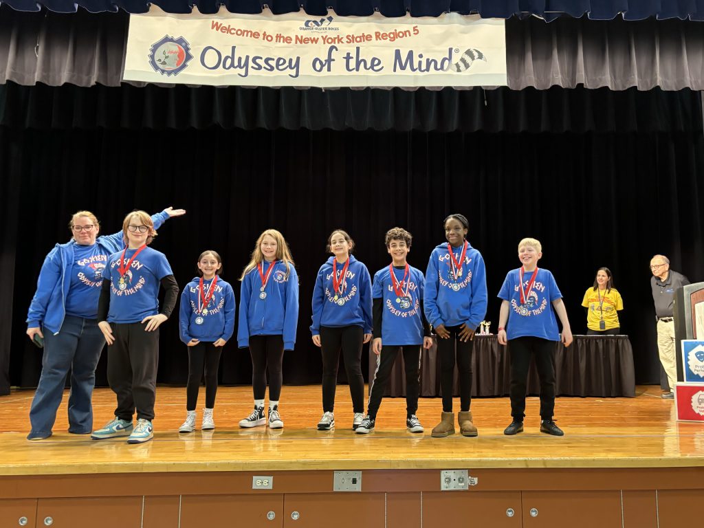 A photo of a winning Odyssey of the Mind team from C.J. Hooker Middle School, proudly wearing medals on a stage under a sign that says "Welcome to the New York State Region 5 Odyssey of the Mind."