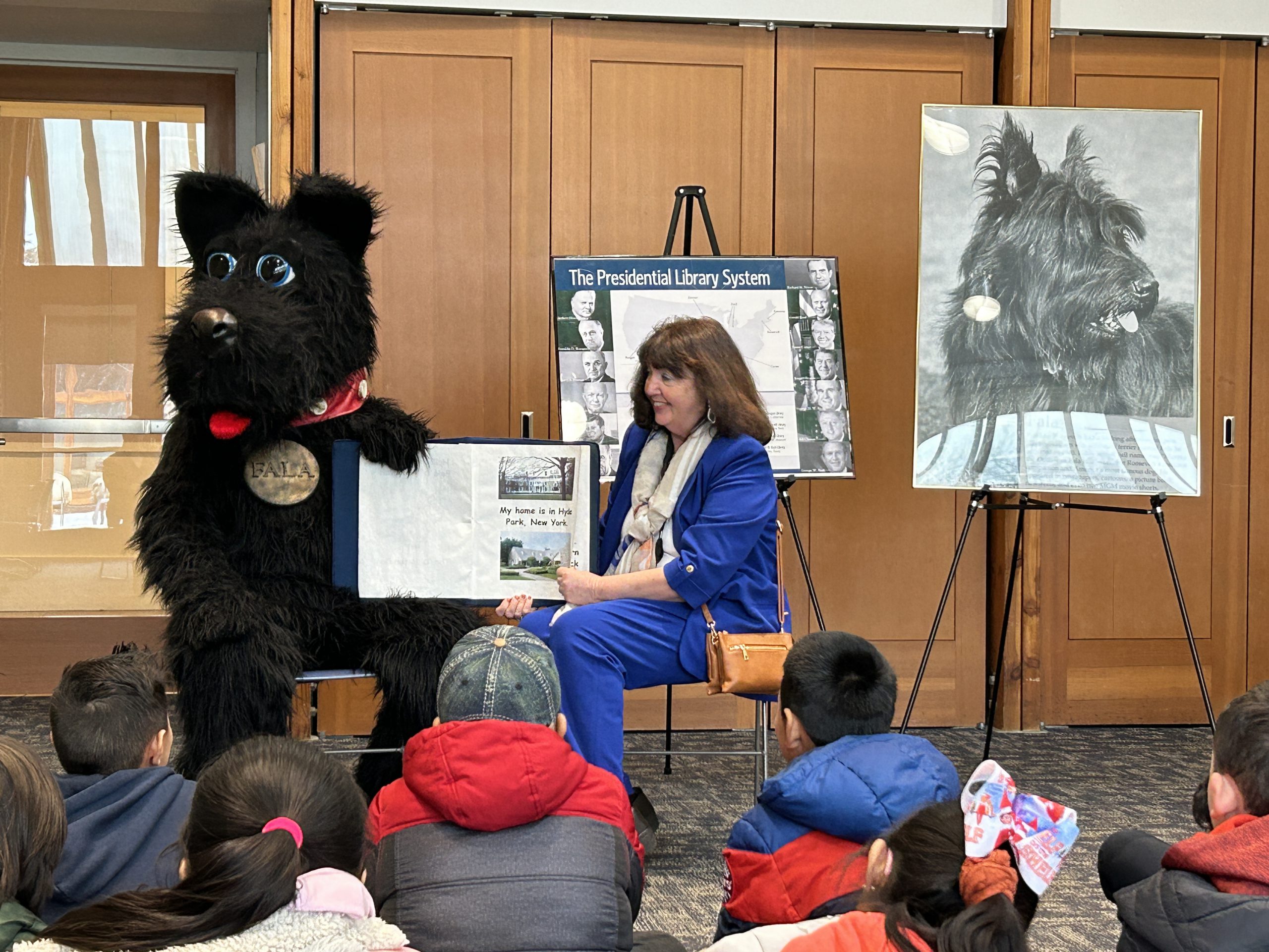 An adult reads from a book next to a person in a black dog costume with a nametag that says "Fala." Intermediate school students look on.