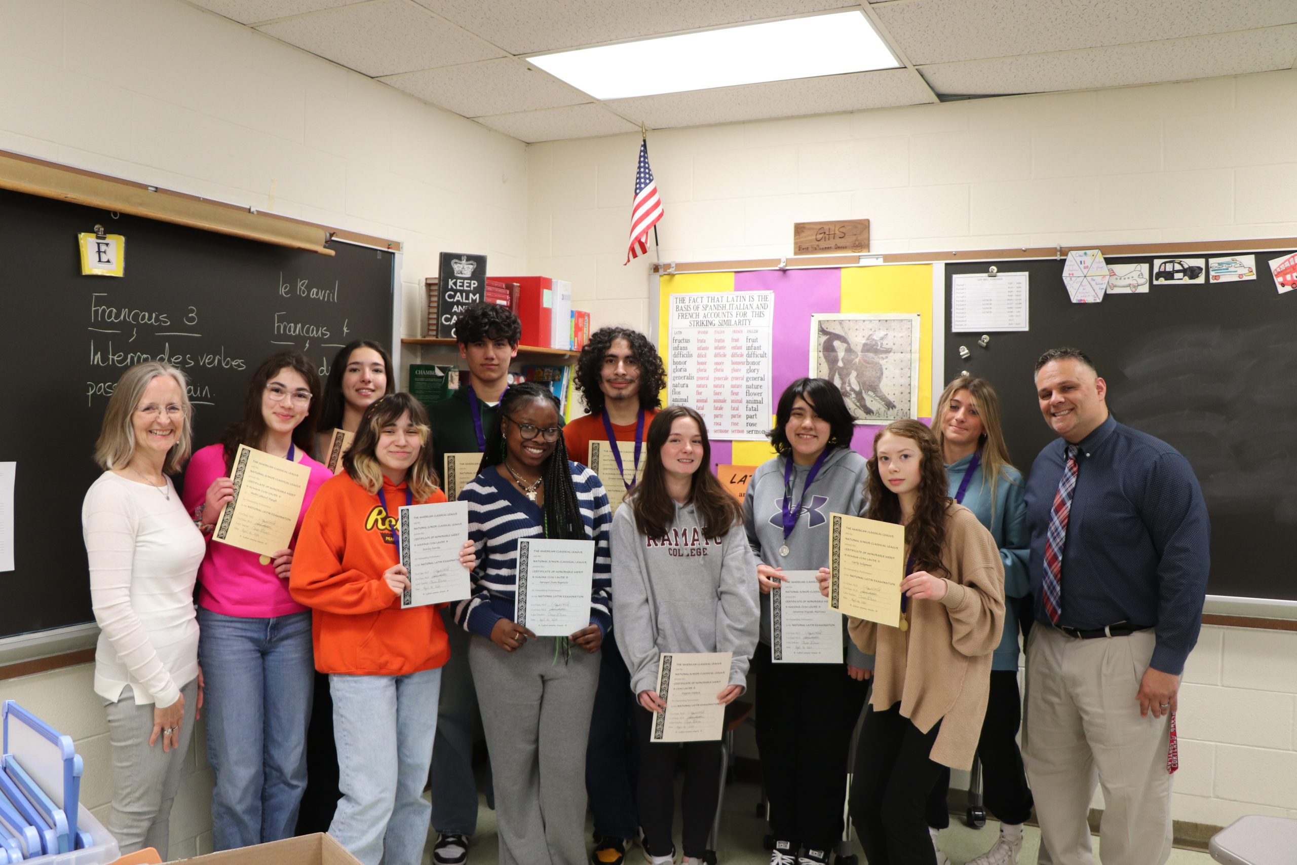 A group of high school students stand holding certificates and wearing medals with two adults in a classroom with posters and writing in Latin and French behind them.