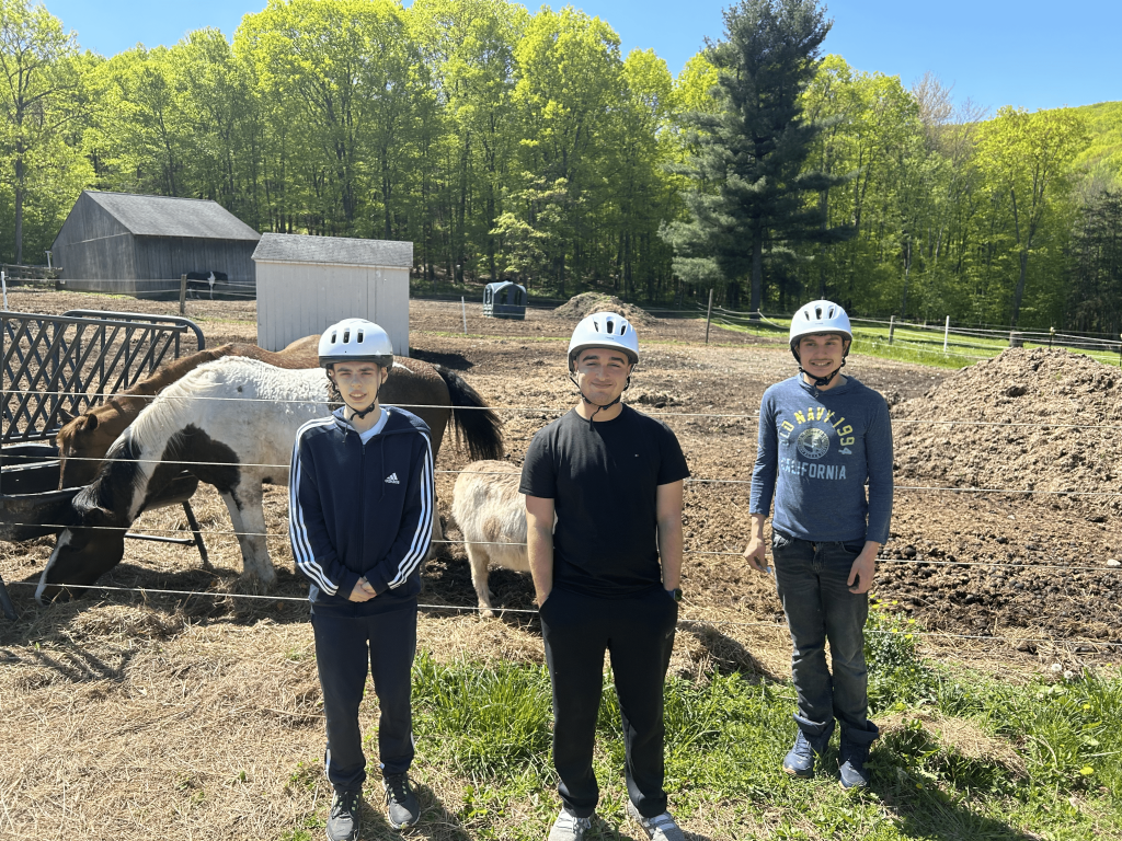 Three teenagers in helmets smile standing in front of horses eating hay in an enclosure outdoors.