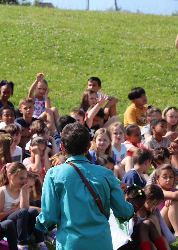 An adult in a teal shirt stands in the foreground with her back facing the camera in front of students sitting on a hill outside with their hands raised to ask questions.