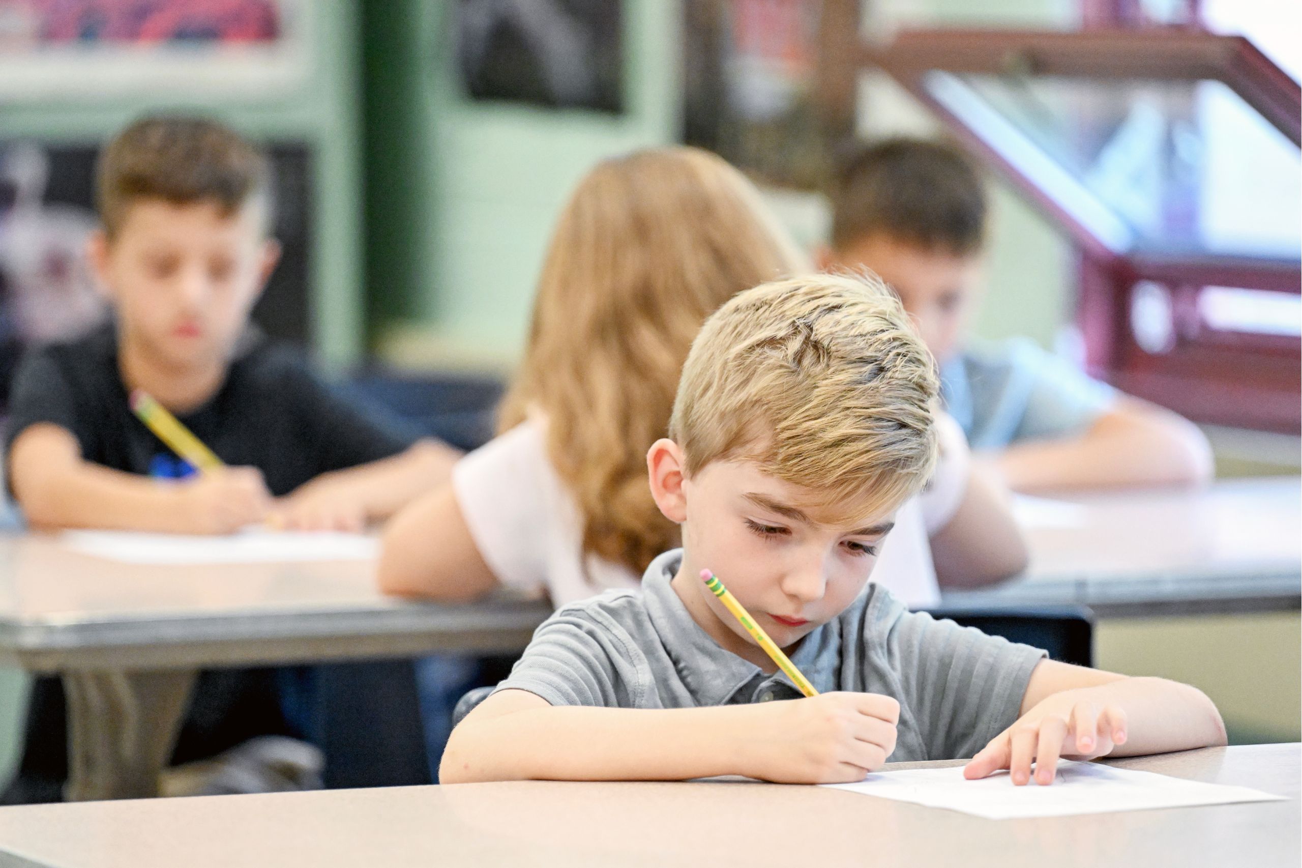 A student is focused on their classwork, writing with a pencil in the air with more students behind them are also focused on class work. 