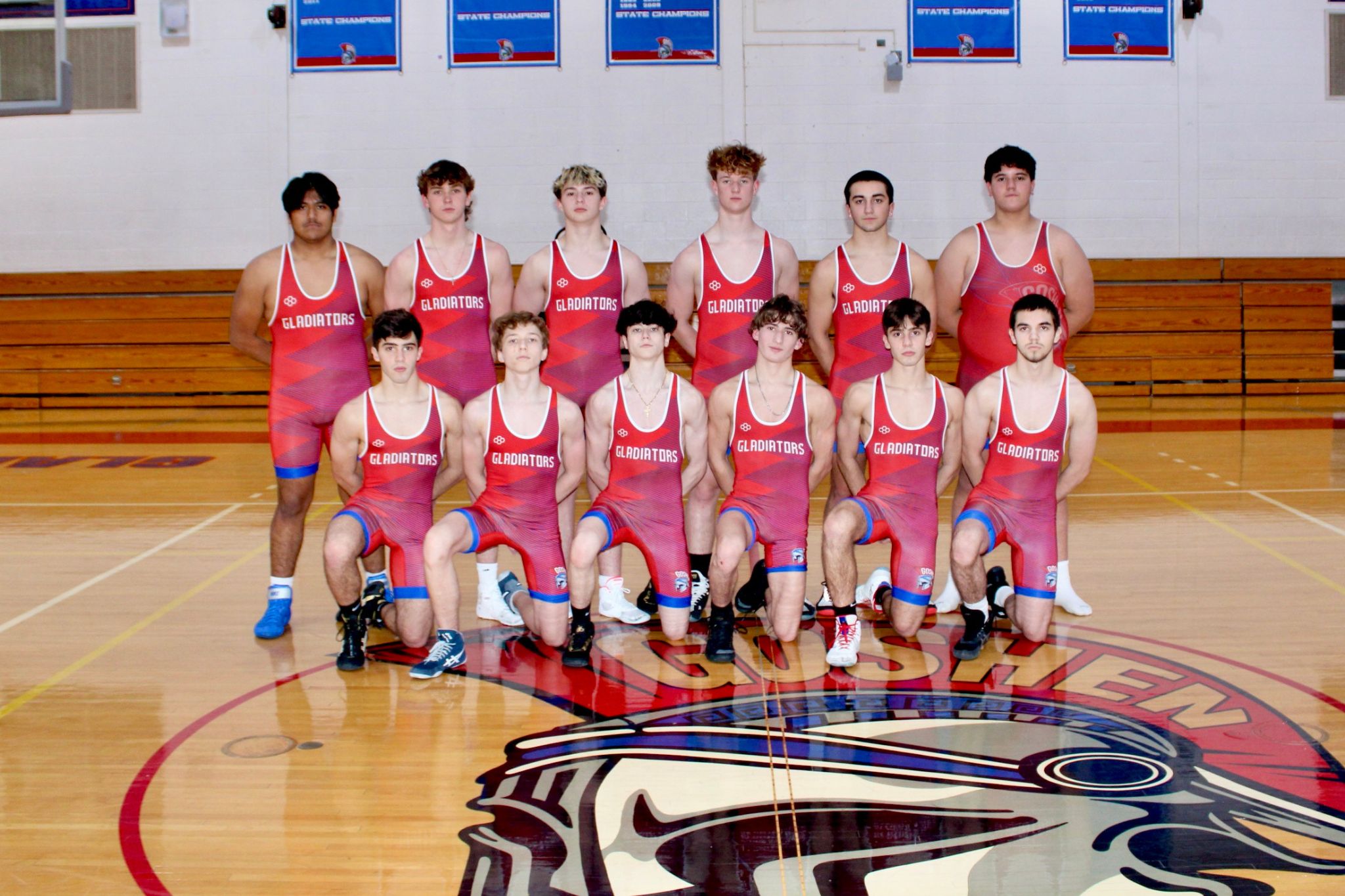 The Goshen Varsity Wrestling team stands together in the gym. The Goshen logo is on the floor. The players are wearing red uniforms with "Gladiators" written across the top. 