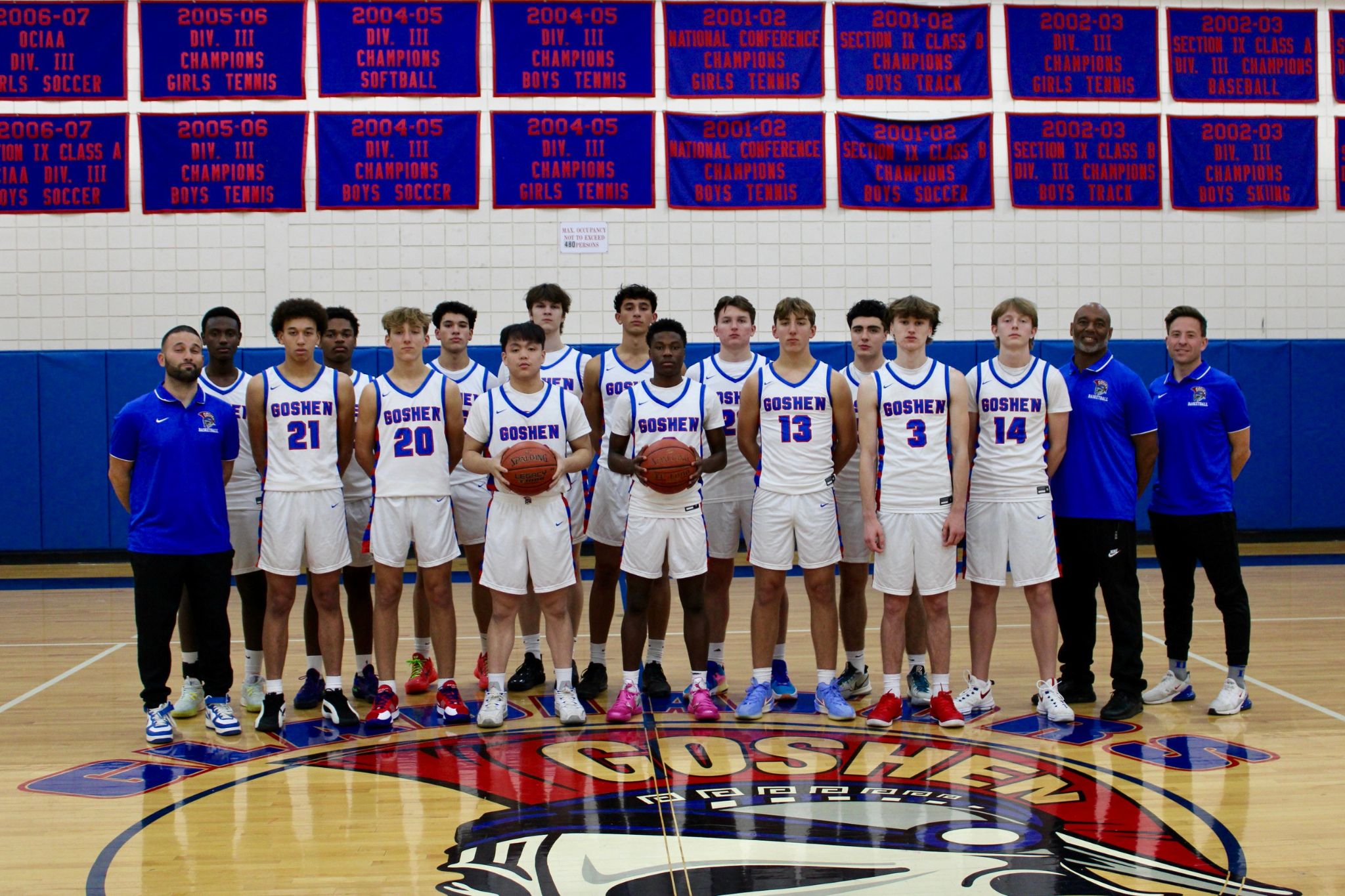 Goshen Varsity Boys basketball team stands for their team photo in the gym. The athletes are wearing their white uniforms with "Goshen" across the jersey. 