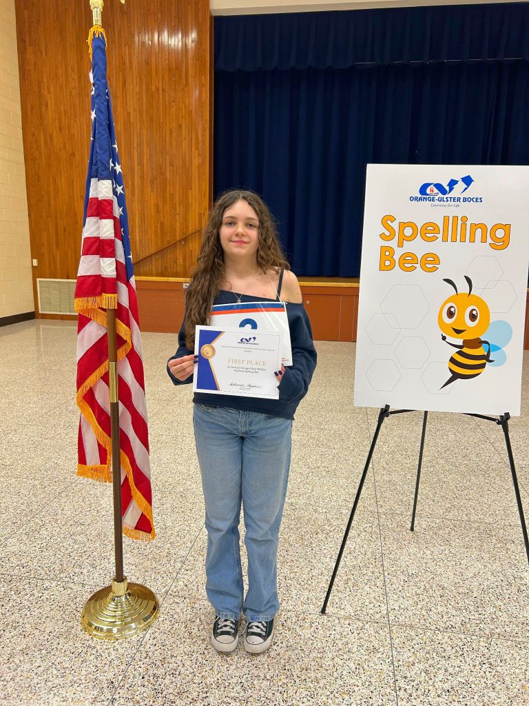 Cora Massaro stands next to the American flag and displays her certificate for winning the Orange-Ulster spelling bee.