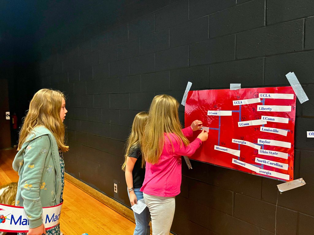 Three girls put their team name in the championship spot on the bracket on the wall.