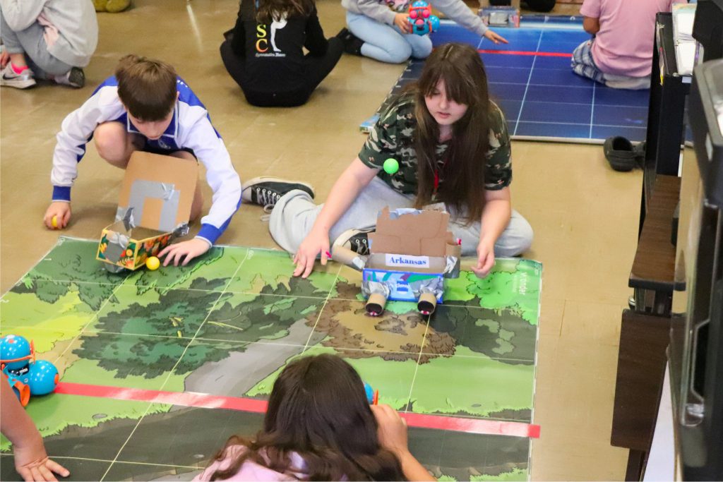 Two teams of students compete to see who can make more baskets in two minutes. The students facing the camera are rebounding the shots Dash is taking and the students on the other end of the mat are refilling Dash to be able to shoot.