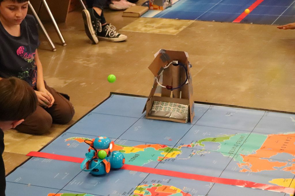 Dash the robot shoots at a student made basket. The basket has pipe cleaners organized into a circle with a cardboard wall holding it up behind the hoop.