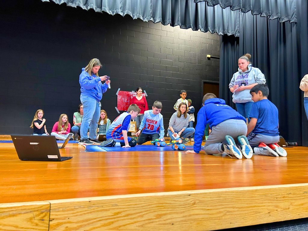 Students on stage are competing for the March Madness championship as they sit on stage near Dash the robot and their mat.