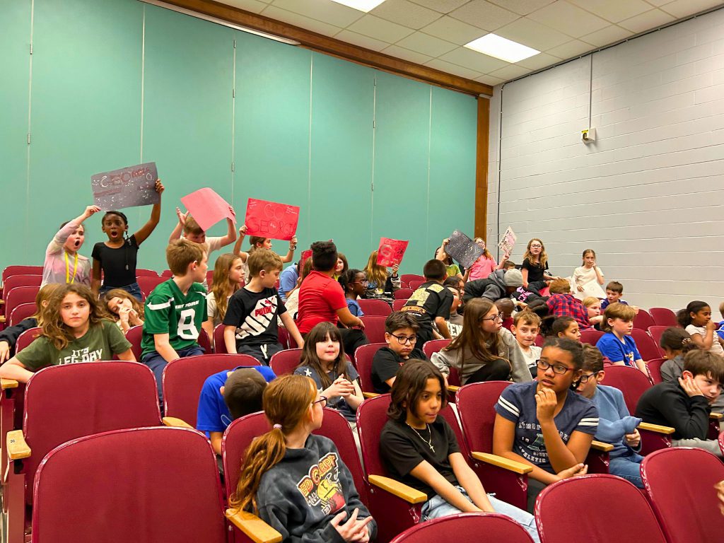 A crowd of students is watching the stage and holding up signs as they cheer on their classmates who are competing for the championship.
