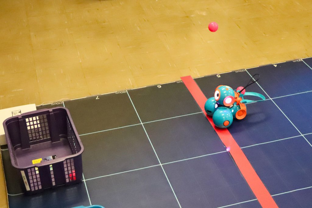 Dash the robot shoots a ball at a plastic container as students practice coding Dash to make shots.
