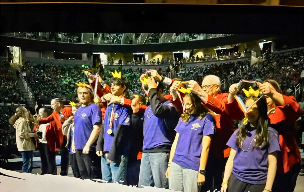 Team Seligman receives their medals from the judges at the Odyssey of the Mind World Finals. They are smiling as medals are placed over their heads.
