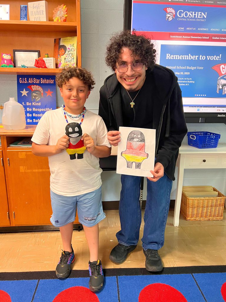 A third grade student stands to the left of a high school student. The young student is holding the sewn monster from the drawing that the older student is holding. The monster has a black head and a red torso with yellow shorts.
