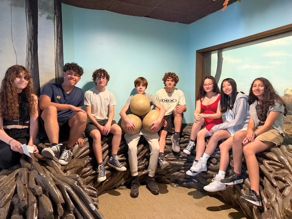 A group of students sit in a room with dinosaur eggs in a large display of a nest. They are sitting in a half circle and looking at the camera.
