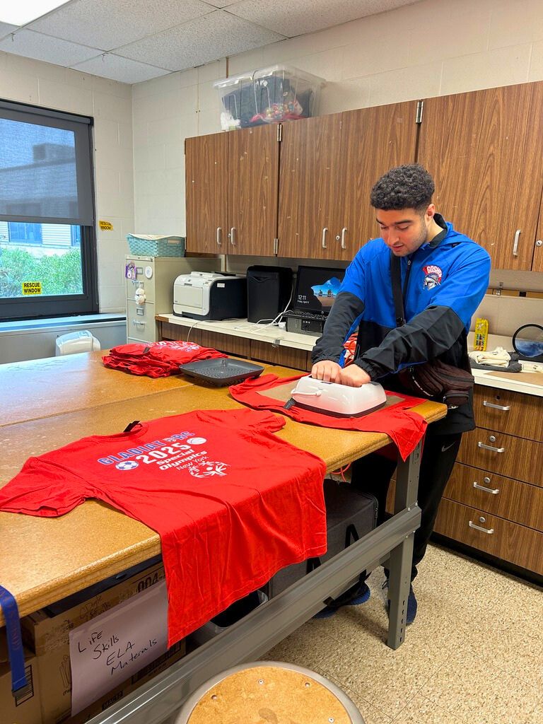 A student holds a machine onto a t-shirt on a table. There is a 2025 Special Olympics shirt on the table next to him facing the camera.