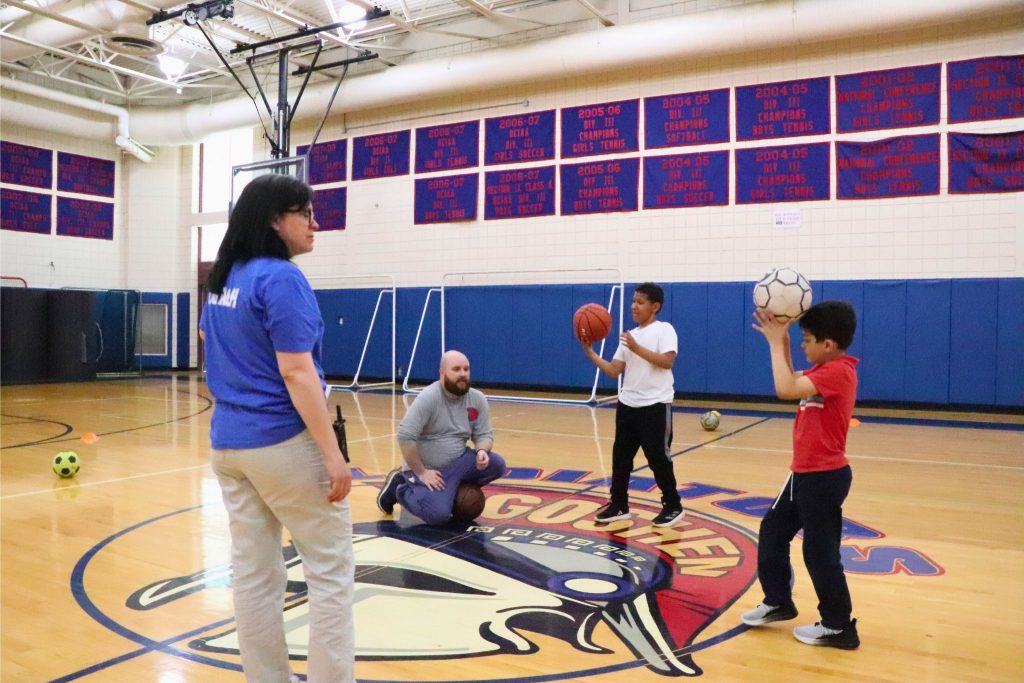 Athletes participating in the upcoming Special Olympics are standing in center court with Kristen Kurpick and Charles Doyle, the coaches of the event. The students have a basketball and a soccer ball.