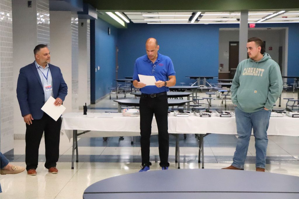 A student stands to the right while a teacher stands and reads in the middle and the Principal, Mr. Pantaleone, stands to the left in the cafeteria.