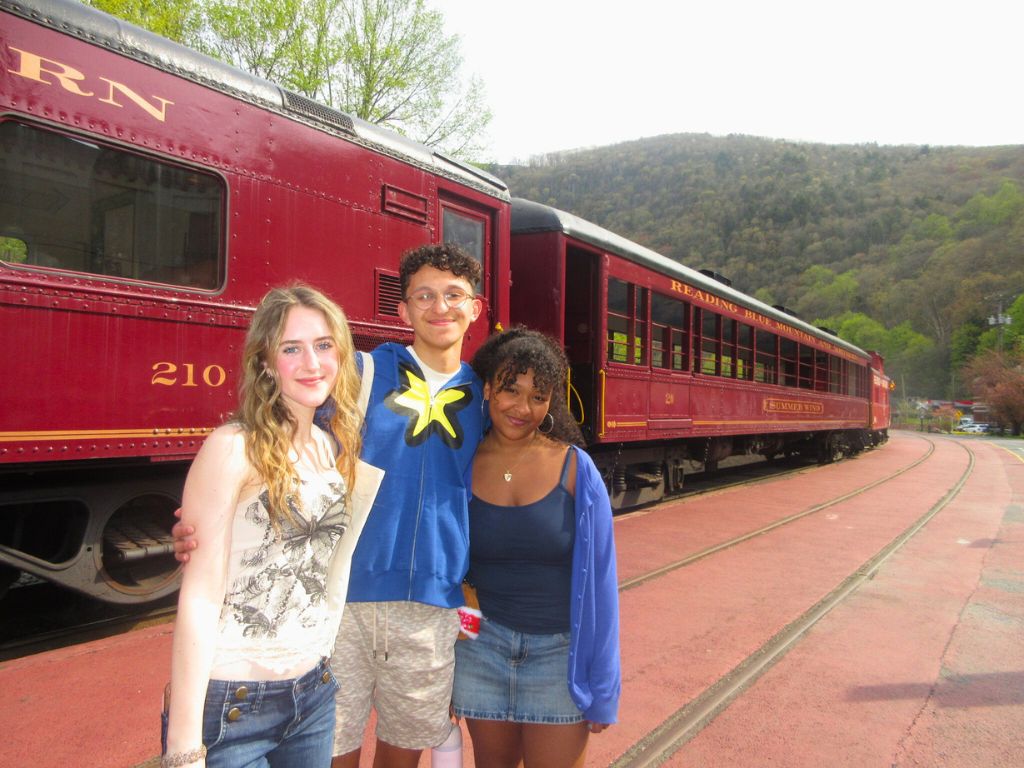Three students are standing next to each other and smiling for a photo in front of a train.