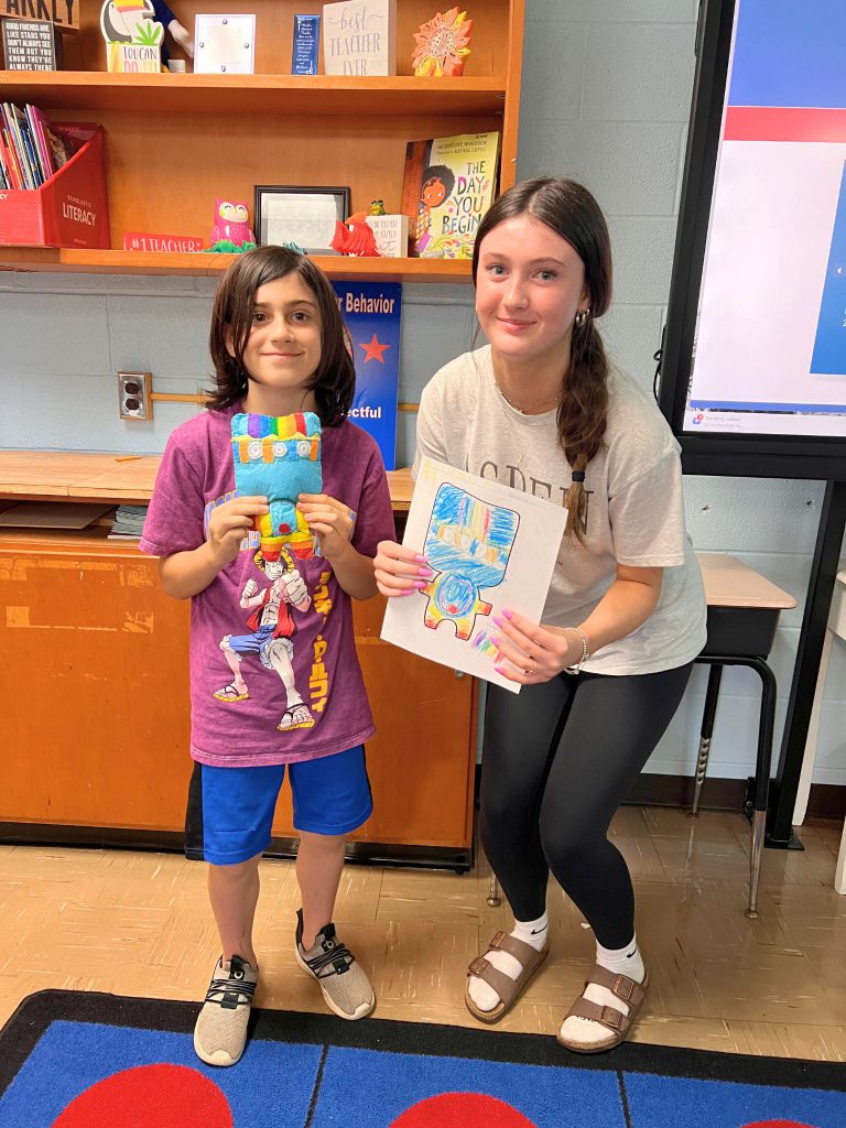 A third grade student stands to the left of a high school student. The young student is holding the sewn monster from the drawing that the older student is holding. The monster is blue and has many colors on its head and legs.