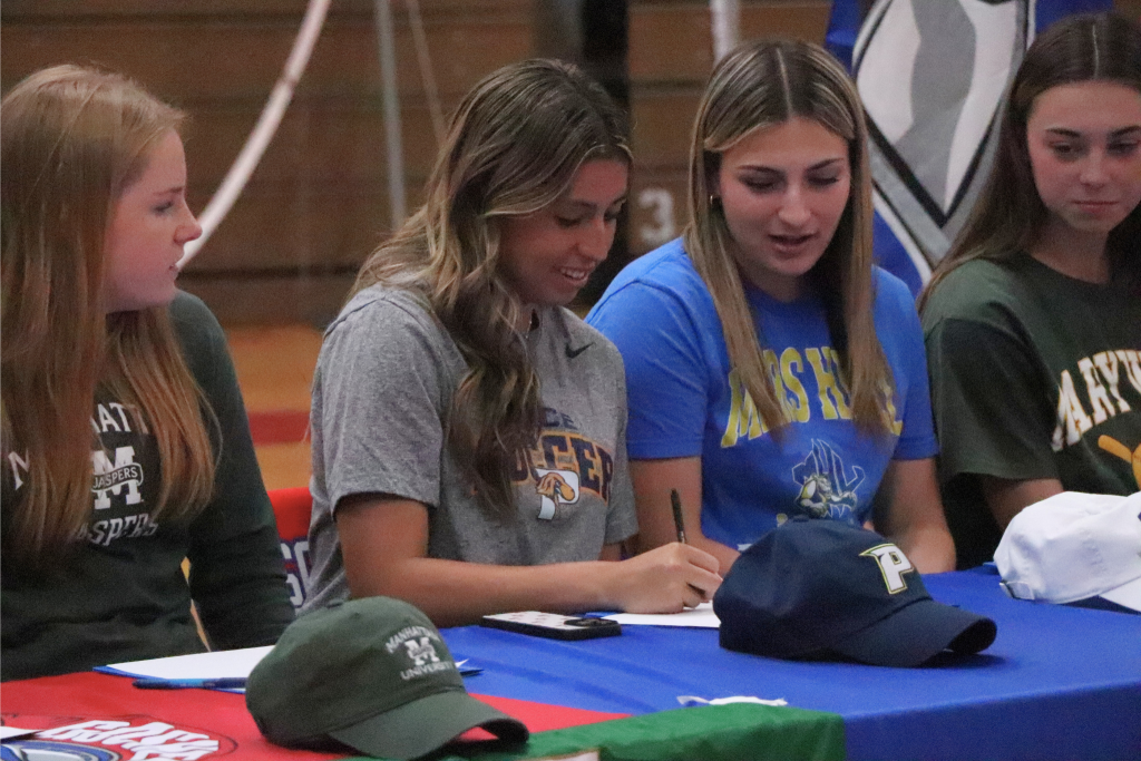 Julia Griffin signs her letter next to Goshen athletes wearing their college's shirt and logo. There is a Pace hat in front of her.
