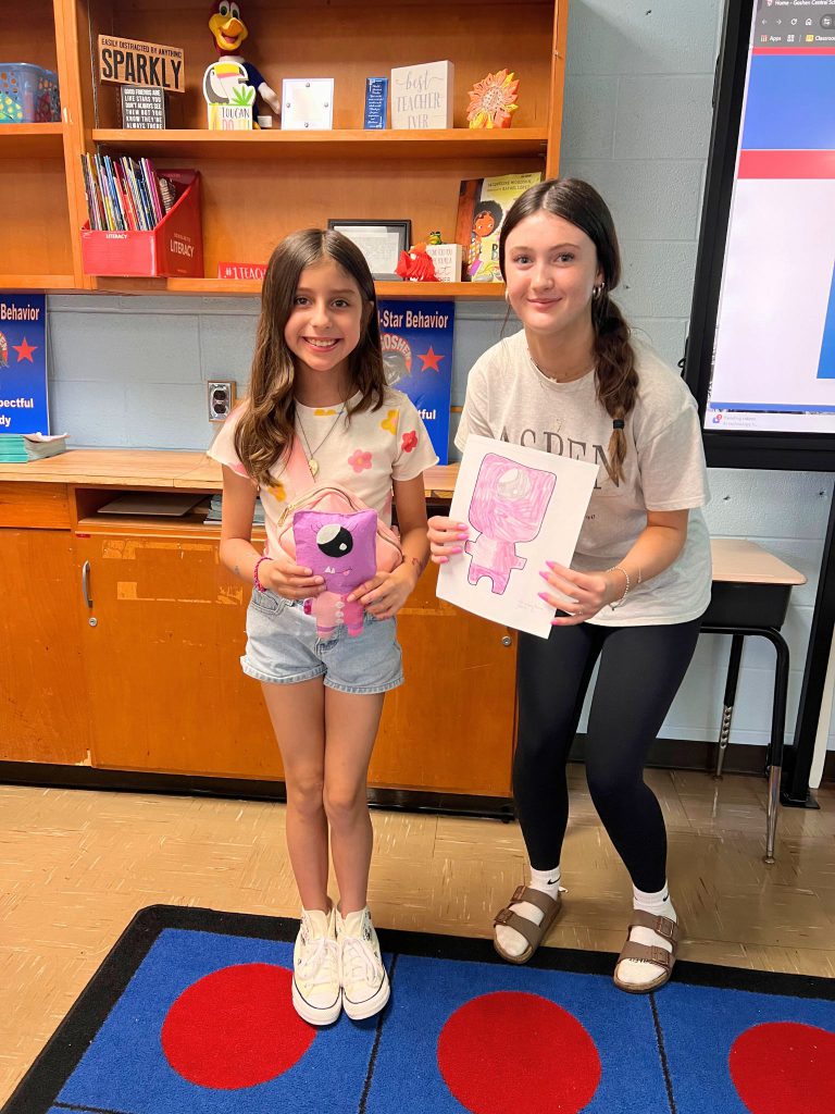 A third grade student stands to the left of a high school student. The young student is holding the sewn monster from the drawing that the older student is holding. The monster is pink and has a big eye in the middle of its head.