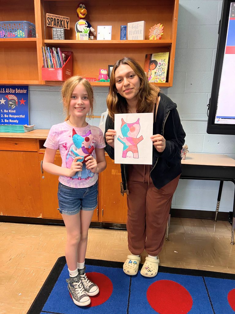 A third grade student stands to the left of a high school student. The young student is holding the sewn monster from the drawing that the older student is holding. The monster is multi-colored with a blue, red, and pink on its body.