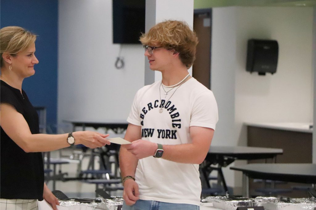 A student looks to the left and accepts a certificate from a teacher in the cafeteria.