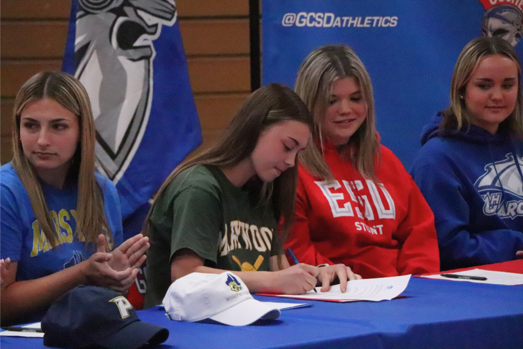 Mia Antolino is signing a letter to make her official commitment to Marywood University to play softball. There are athletes around her clapping as she signs.
