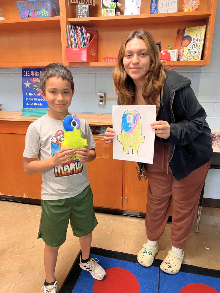 A third grade student stands to the left of a high school student. The young student is holding the sewn monster from the drawing that the older student is holding. The monster is yellow with a blue and purple head and an eye in the middle of its head.