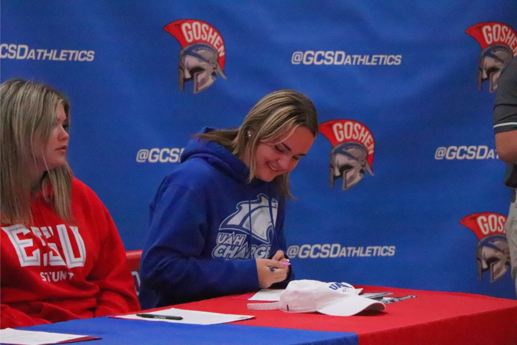 Meghan Doody signs her letter to play lacrosse at the University of Alabama Huntsville. She is smiling and sitting in front of a Goshen background with her college's hat in front of her.
