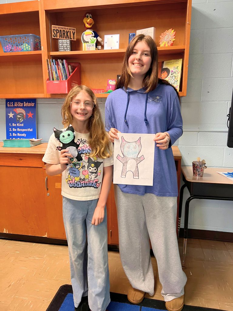 A third grade student stands to the left of a high school student. The young student is holding the sewn monster from the drawing that the older student is holding. The monster is black but has a white belly and a turquoise face.