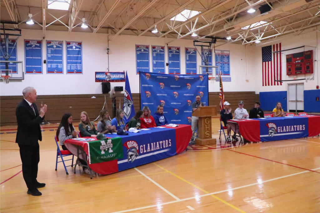 Jarrad Vrydaghs and George St. Lawrence stand and clap for the Goshen athletes that are attending Sports Signing Day. They are sitting at a Goshen Gladiators table on either side of the GHS podium.