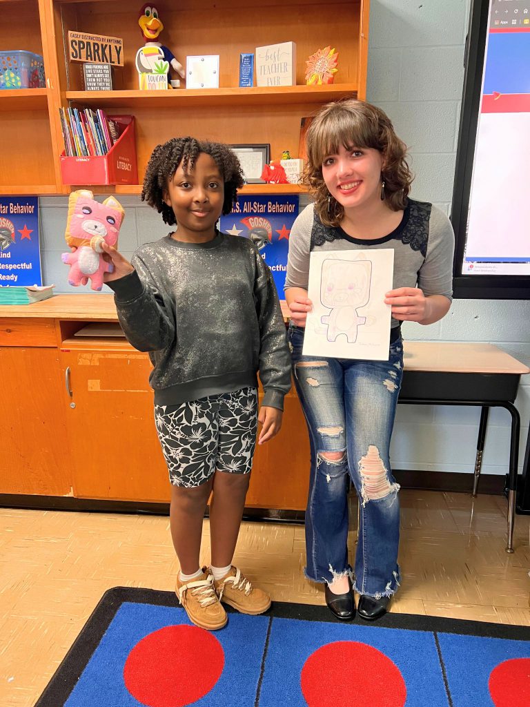 A third grade student stands to the left of a high school student. The young student is holding the sewn monster from the drawing that the older student is holding. The monster is pink and has white on its nose and stomach and has ears as well.