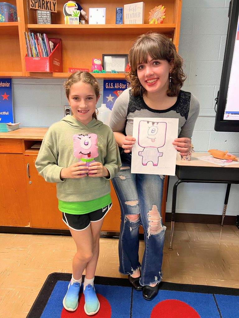 A third grade student stands to the left of a high school student. The young student is holding the sewn monster from the drawing that the older student is holding. The monster is pink with a big eye in the middle of its head and has its tongue sticking out.