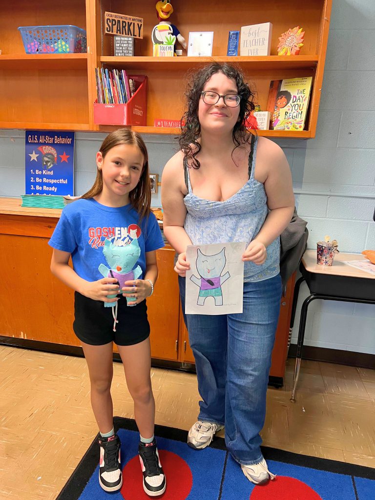A third grade student stands to the left of a high school student. The young student is holding the sewn monster from the drawing that the older student is holding. The monster is light blue with a purple shirt and light blue pants.