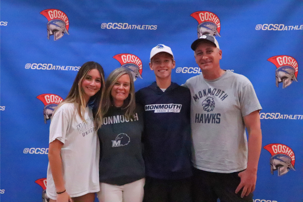 Noah Klugman and his family are smiling and looking at the camera in front of a Goshen Gladiators background. They are all wearing Monmouth gear.