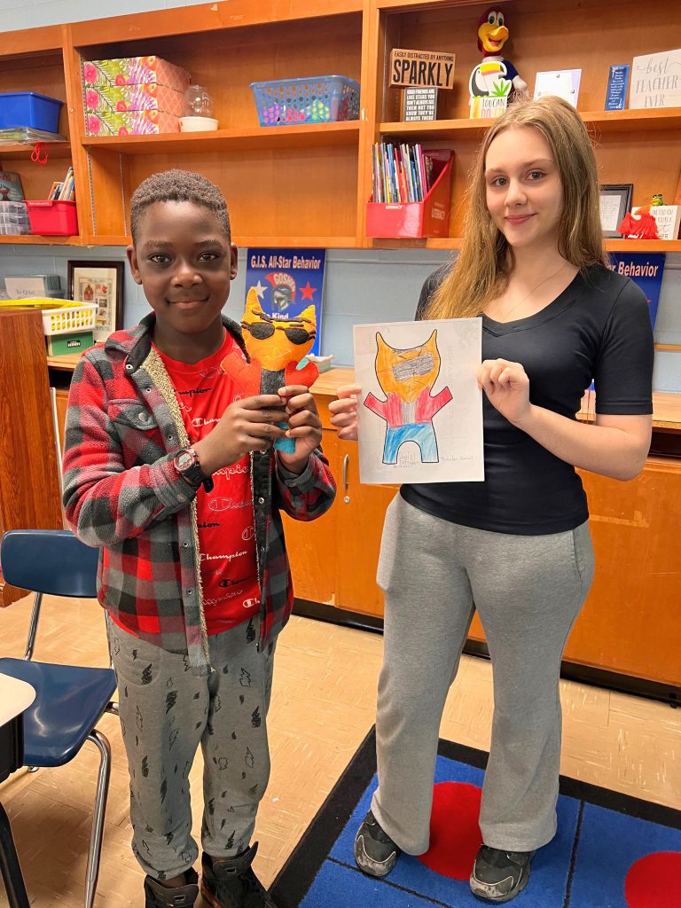 A third grade student stands to the left of a high school student. The young student is holding the sewn monster from the drawing that the older student is holding. The monster is orange with a red shirt on and blue pants, along with sunglasses.