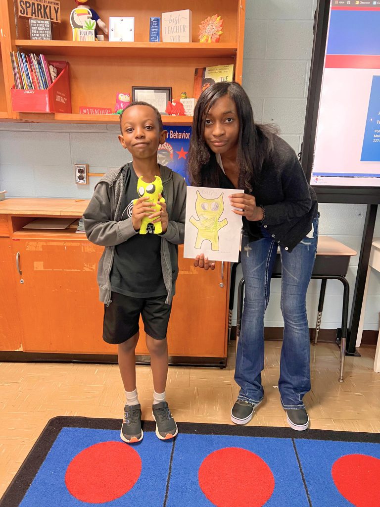 A third grade student stands to the left of a high school student. The young student is holding the sewn monster from the drawing that the older student is holding. The monster is yellow and has horns-like points coming up off the top of it's head.