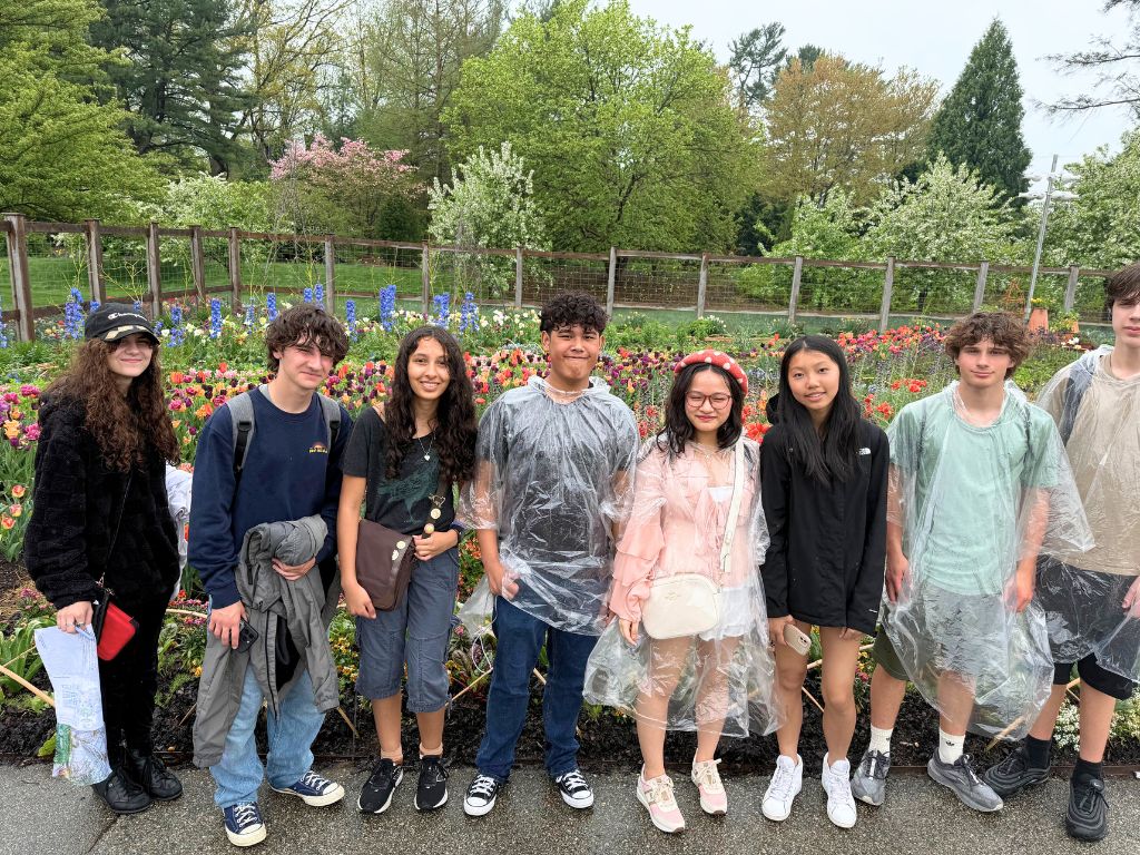 Eight students are wearing rain ponchos and are smiling at the camera in front of a garden.