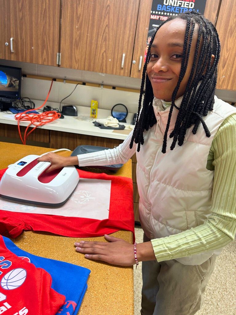 A student smiles for the camera while holding a white machine that is pressing the Special Olympics logo onto the shirt on the table.