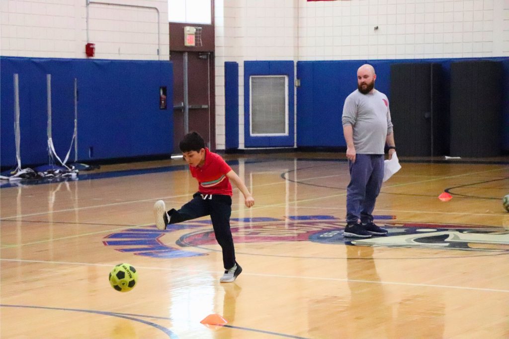 A student kicks a soccer ball as coach Charles Doyle watches behind him.