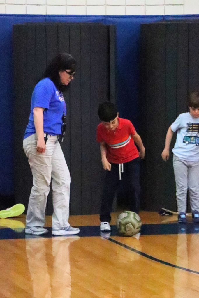 Coach Kristen Kurpick stands next to two students practicing for the Elementary Special Olympics. They have a soccer ball near the wall in the gym.