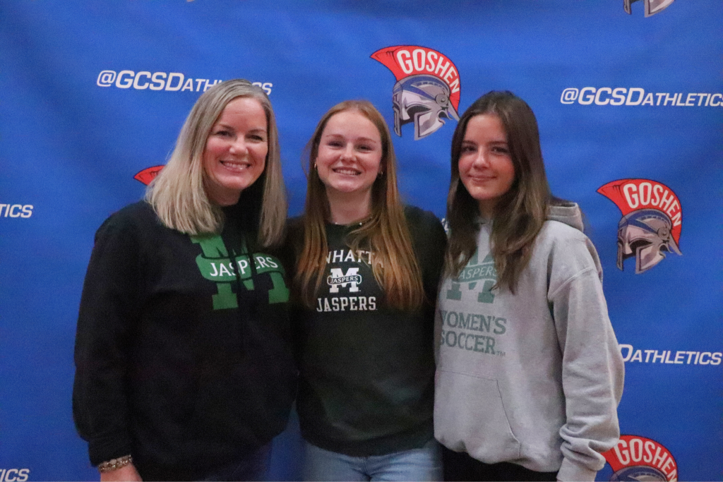 Emma Duffy stands and smiles with her family. They are in front of a Goshen background and wearing Manhattan sweaters.