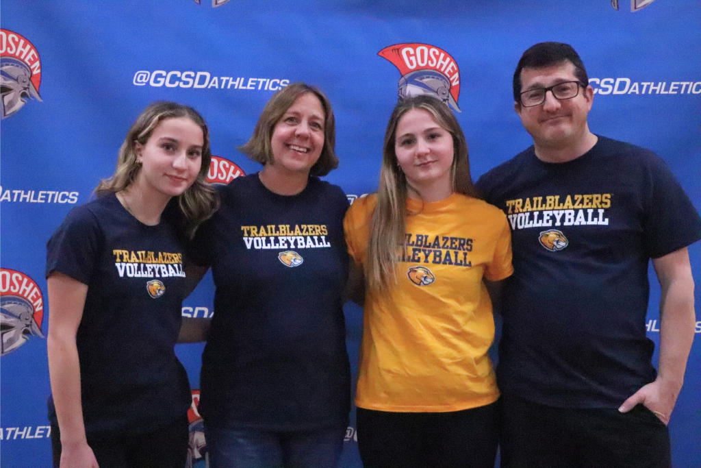 Olivia Grob stands in front of a Goshen background and smiles with her family. They are all wearing Trailblazers volleyball shirts and the logo.