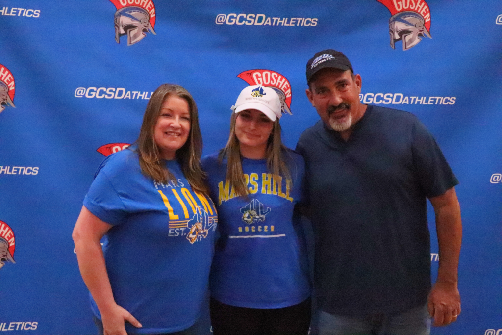 Sophia Appello smiles with her family with the Goshen Gladiators background. They are wearing Mars Hill hats and shirts.