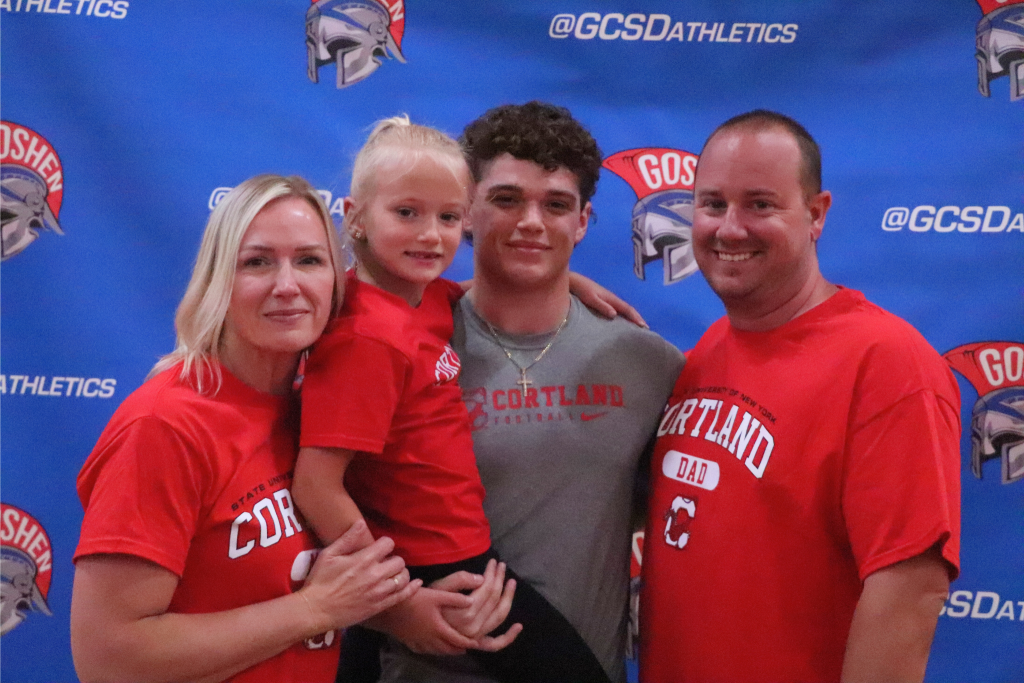 Vincenzo Pupo smiles with his family in front of a Goshen background. They are all wearing Cortland shirts, including a child Vincent is holding. A family member is on either side of him.