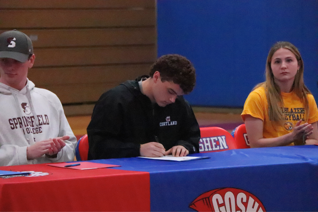 Vincenzo Pupo signs a paper to make official his attendance at SUNY Cortland to play football. Two Goshen athletes sit next to him and clap at a table decorated in Goshen logos.