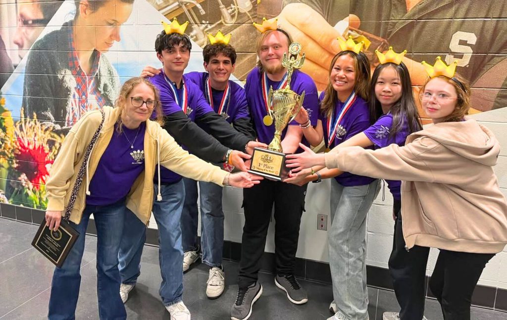 Team Seligman holds up their 1st place trophy. The team is wearing crowns on their heads and smiles for a photo.