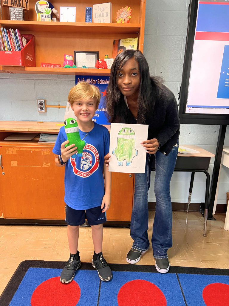 A third grade student stands to the left of a high school student. The young student is holding the sewn monster from the drawing that the older student is holding. The monster is light green and has big teeth it is showing at all times.